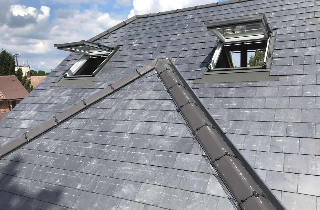 Roof featuring natural slate tiles with two open skylights, showcasing a pitched design and a ridge cap, under a partly cloudy sky.