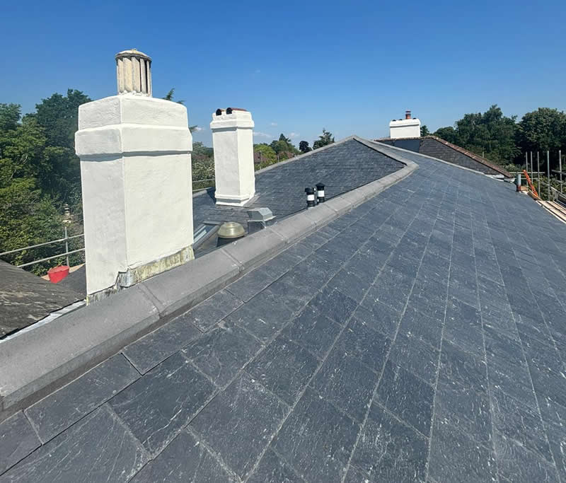 Natural slate roof installation featuring two white chimney stacks, surrounding trees, and clear blue sky, emphasizing craftsmanship and durability for heritage properties.