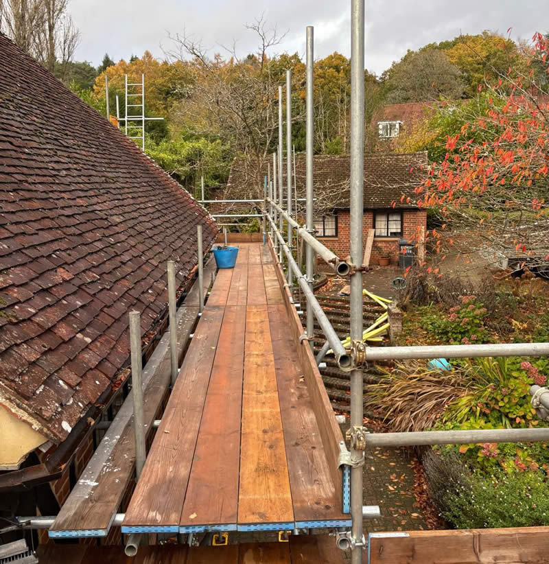 Scaffolding platform installed for roofing work, featuring wooden planks and metal supports, surrounded by a residential garden and trees, illustrating safe access for building restoration projects.
