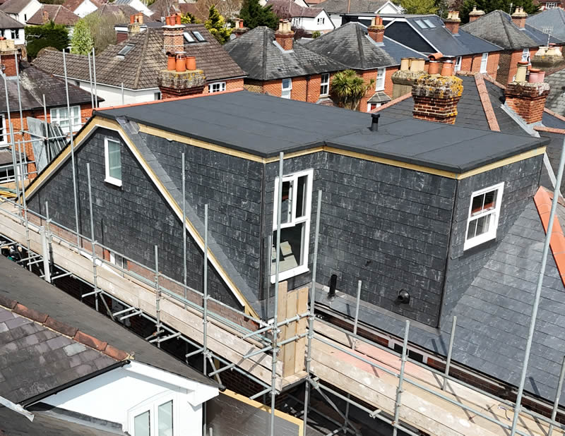 Loft conversion roof under construction with scaffolding, featuring slate tiles and large windows, integrated with existing structures and designed for durability and aesthetics.