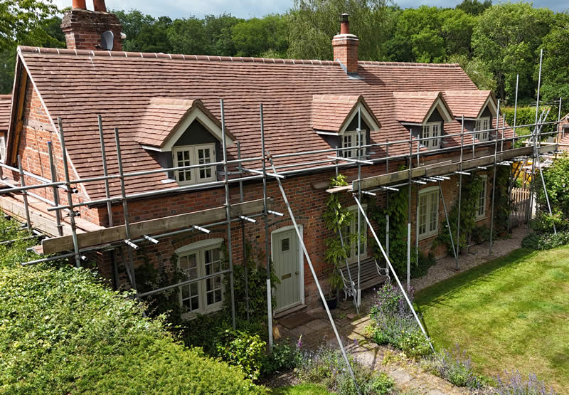 Traditional brick cottage with clay plain tile roof under scaffolding, surrounded by lush greenery and garden, showcasing precision roofing work by specialists.